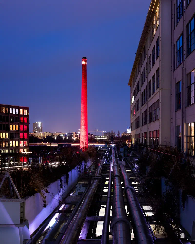 Red-lit chimney of Strijp-S beyond a bundle of pipes running between former industrial Philips buildings at dusk