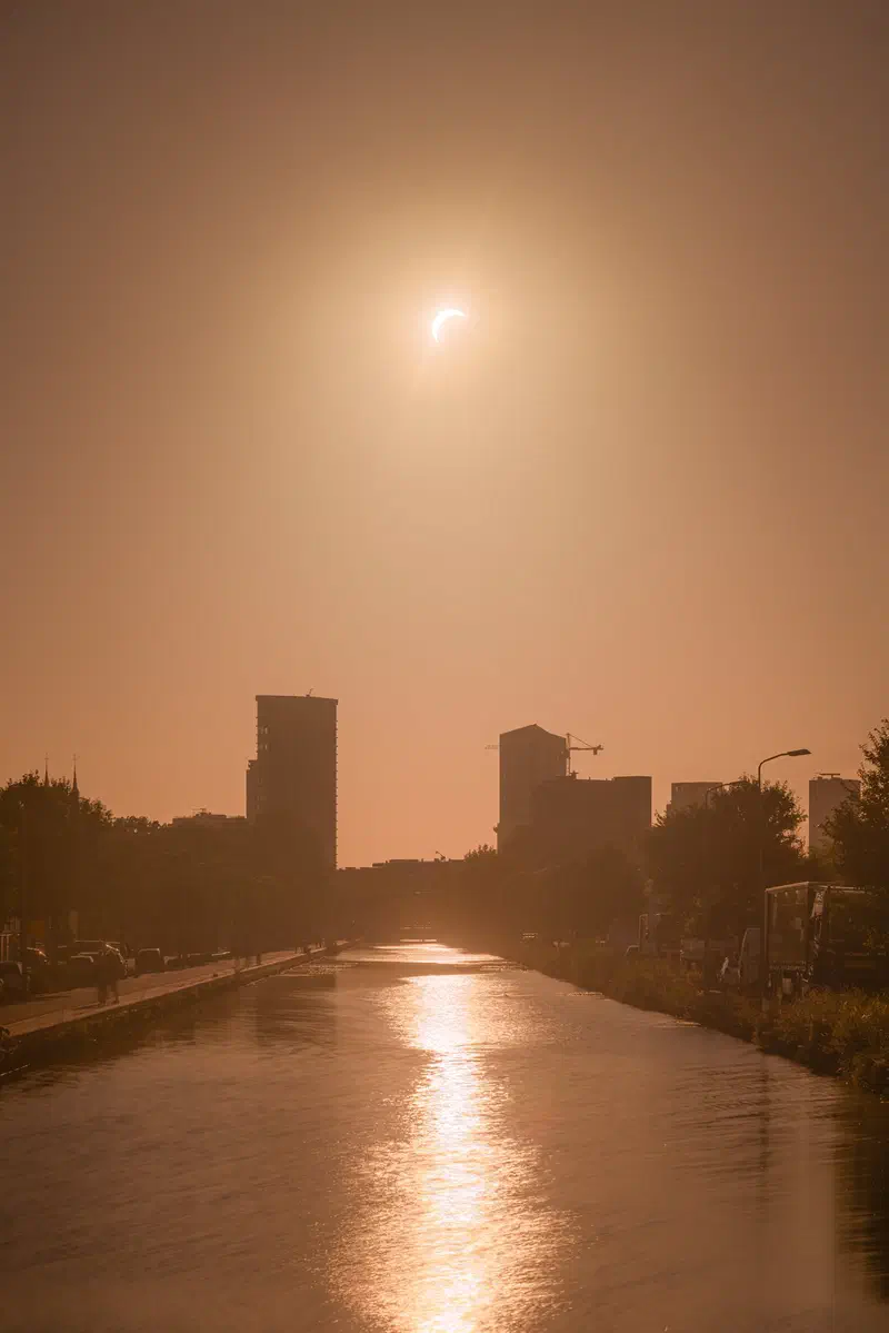 Hazy solar eclipse above a canal (Eindhovens Kanaal) and distant buildings (Vestedatoren & Admirant) in an amber sky
