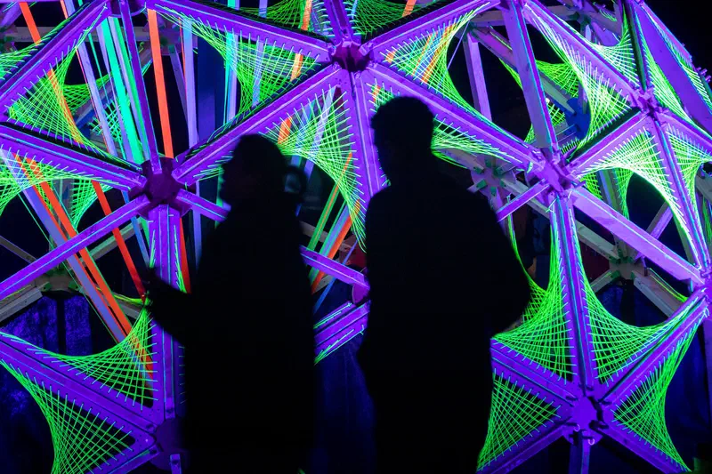 Silhouettes of two people in front of a neon string-art structure (Glow)