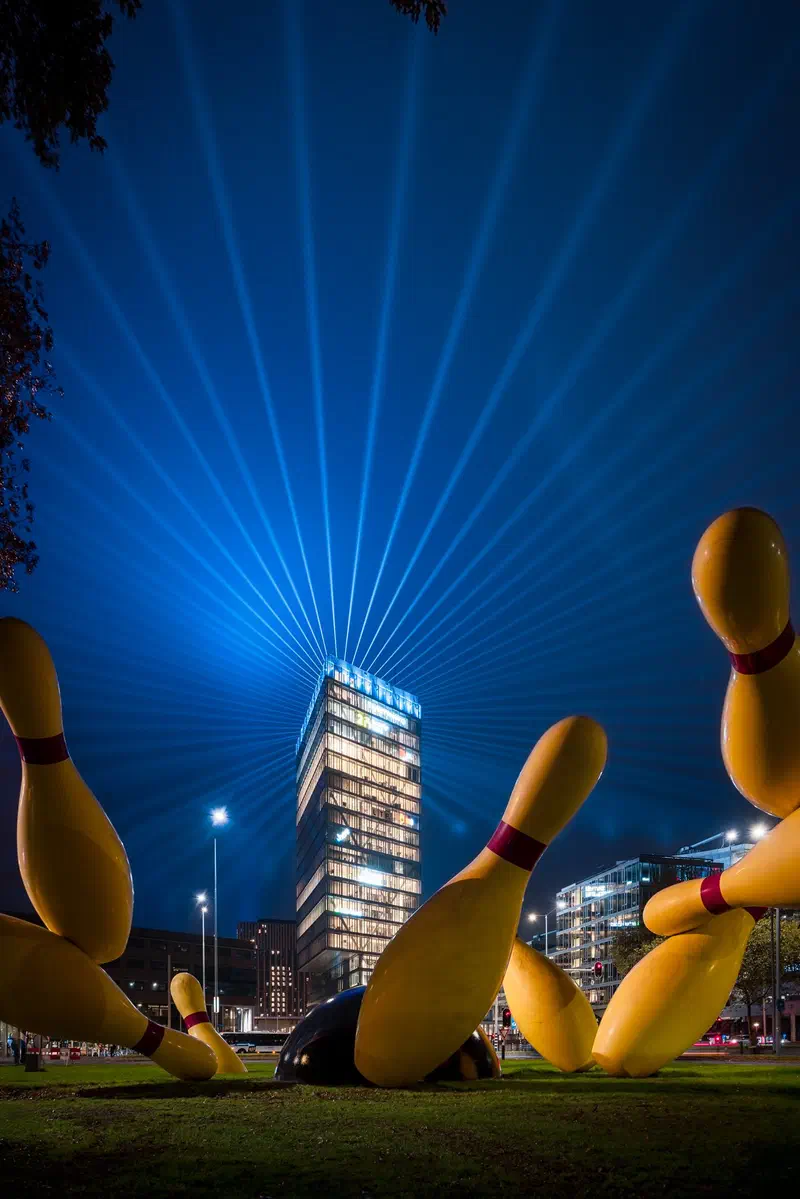 Giant yellow bowling pins (Flying Pins) on a lawn with laser beams (Glow) above a tower (Kennedytoren)