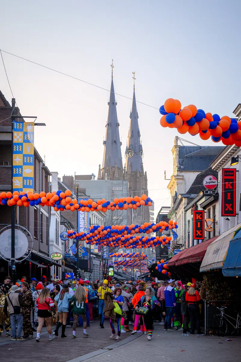 Costumed crowd in a street (Stratumseind) hung with orange and blue balloons below Sint-Catharinakerk during Carnaval