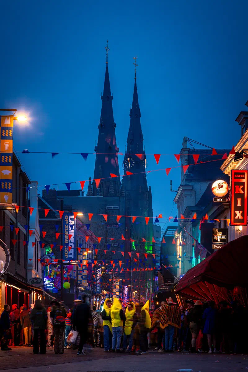 Twin spires of Sint-Catharinakerk at the end of a street (Stratumseind) with bunting and people in yellow ponchos during Carnaval