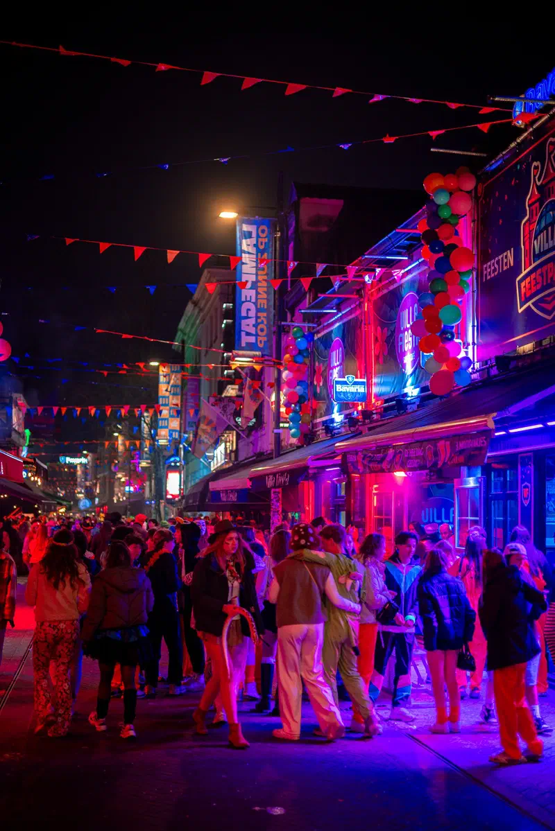 Packed street (Stratumseind) under bunting with pink and purple neon lights at night during Carnaval