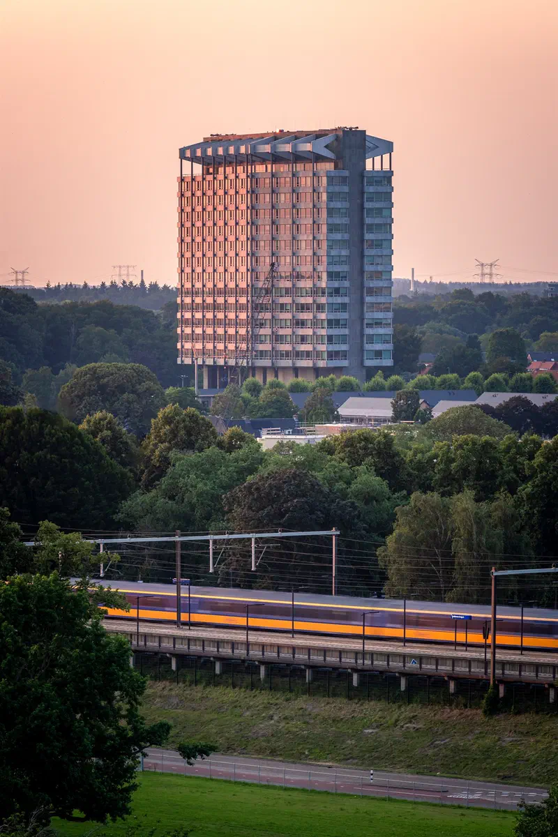 Tall building (Philips Nederland) above green trees with a passing train in warm evening light