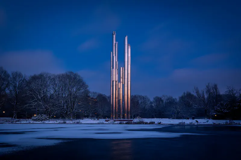 Hoge metalen pilaren (Schoorsteenplastiek) naast een bevroren vijver (Henri Dunantpark) in de schemering
