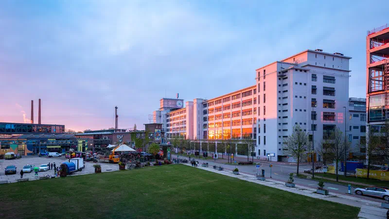 Former factory buildings lit by golden sunrise beyond a green lawn, Strijp-S