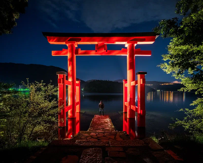 Red torii gate in Japan lit at night, with a lone figure facing a lake